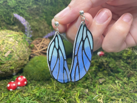 Blue butterfly-wing earrings held against a natural background with moss and small red mushrooms.