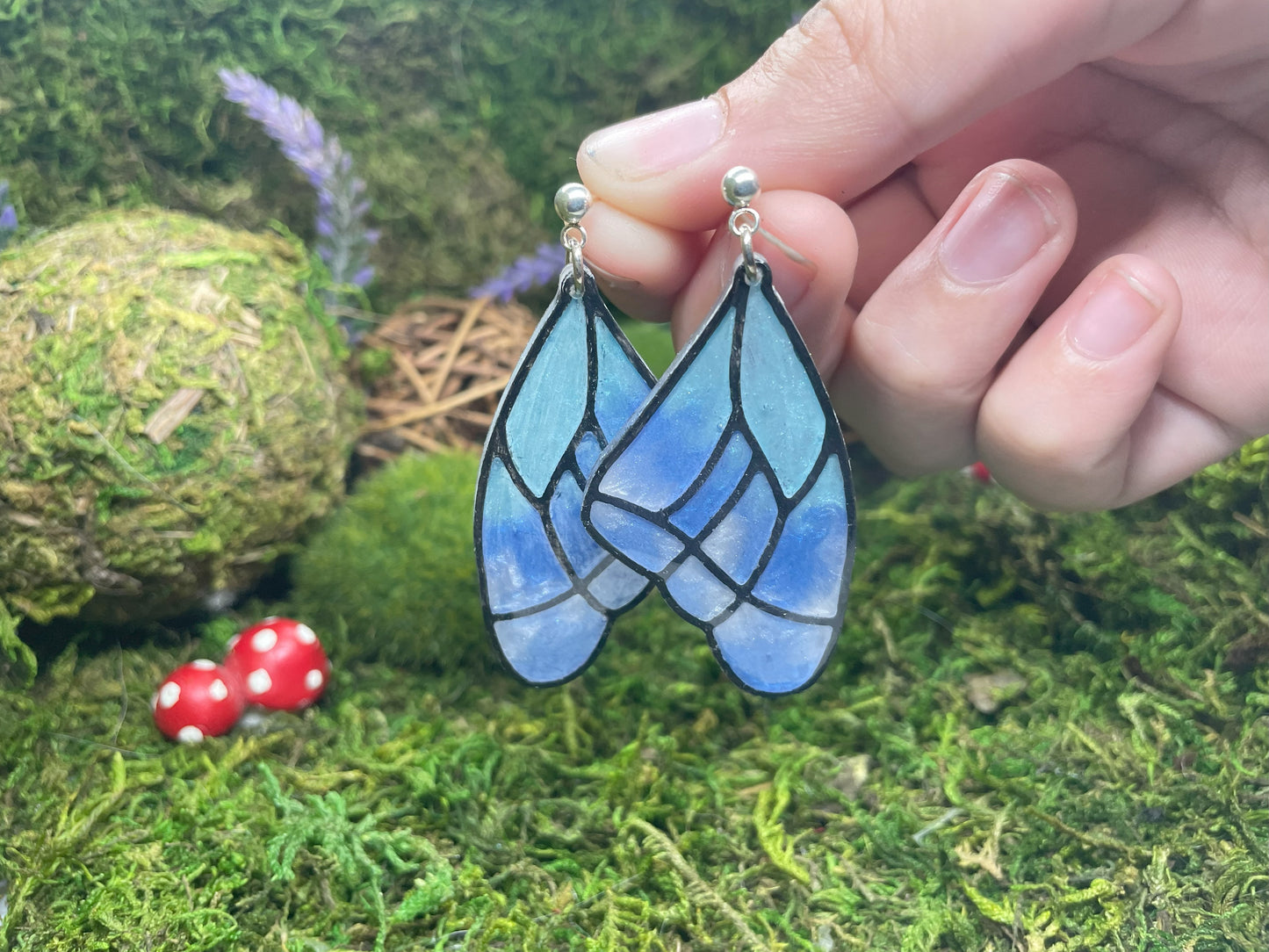 Blue butterfly wing earrings held by a hand against a natural background with moss and small red mushrooms.