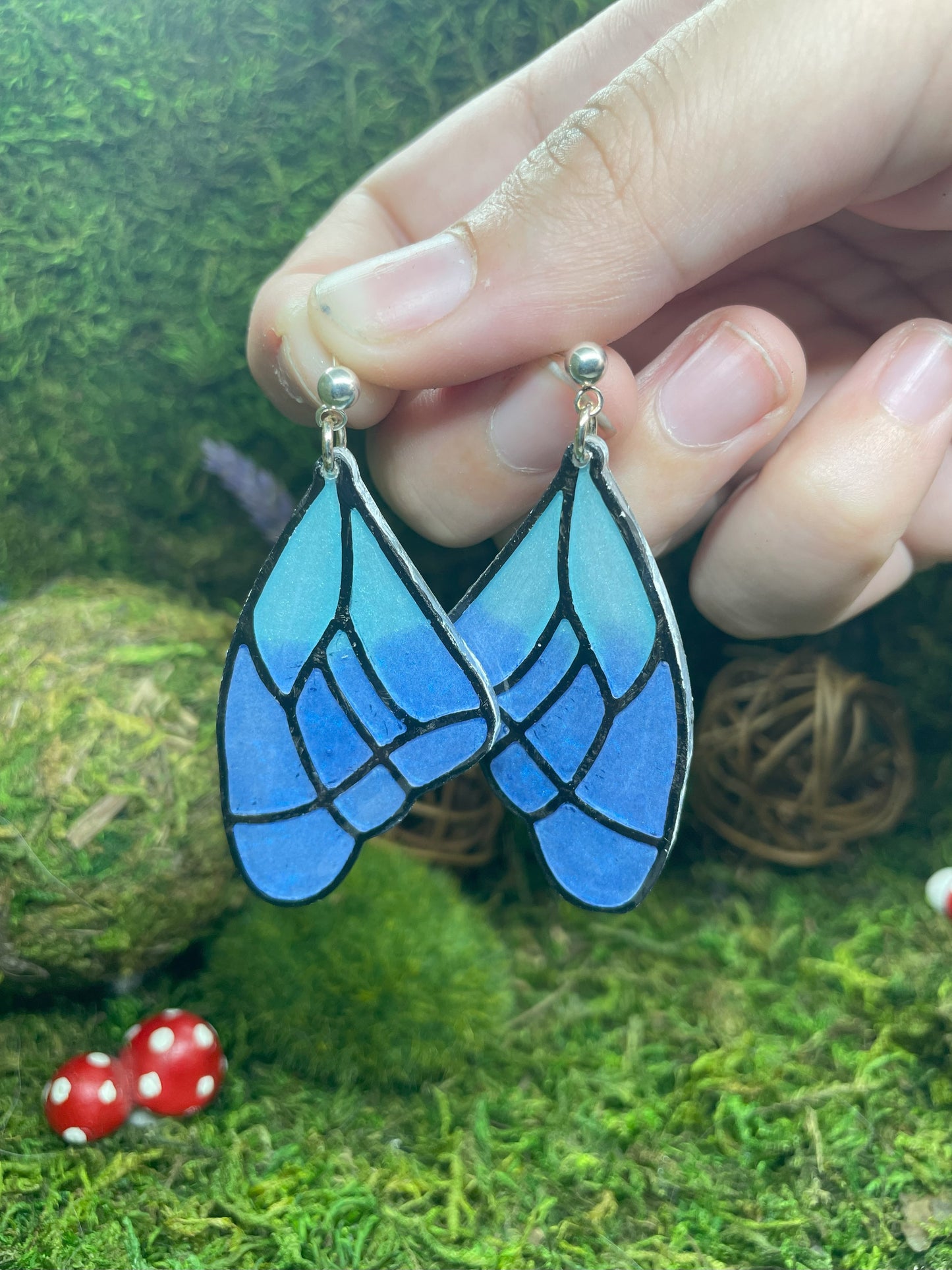 Blue butterfly wing earrings held by a hand on a green moss background with red mushrooms.
