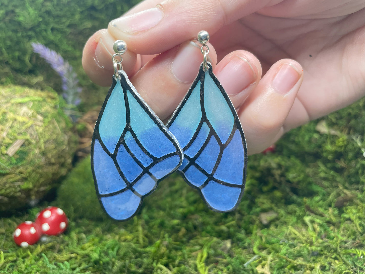 Blue butterfly-shaped earrings held against a natural background with moss and small red mushrooms.