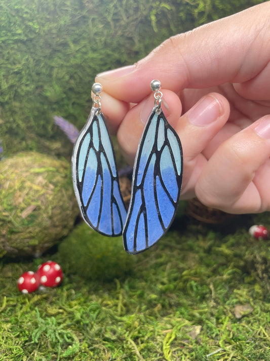Blue butterfly wing earrings held by a hand against a green mossy background